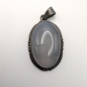 May include: Oval-shaped pendant with a light gray stone set in a silver-toned metal frame. The frame has a decorative border and a loop for a chain. The stone has a translucent quality.