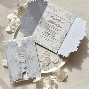Puede incluir: Un conjunto de invitaciones de boda gris claro con intrincados patrones florales cortados con láser en las solapas. El texto de la invitación incluye los nombres Evelyn Marsh y Anthony Pierce, junto a varios sellos de cera circulares.