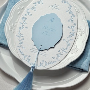 May include: An oval white menu card with a light blue floral border and a smaller, scalloped light blue card attached by a light blue tassel. The menu features the text Menu, the name Charlotte Chase, and the words Vanilla Bean Cake.