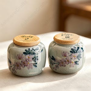 May include: Two ceramic urns with wooden lids. The urns are pale green with a floral design in pink and blue. One lid says "Rocky" and the other says "Kitten". Both lids have dates and text.