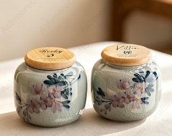 May include: Two ceramic urns with wooden lids. The urns are light green with a floral design in pink and blue. One lid says "Rocky" and the other says "Kitten". Both lids have dates and text.