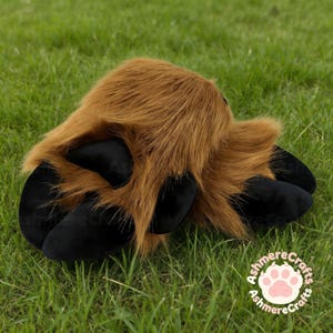 May include: A brown faux fur paw with black claws, lying on green grass. The paw has a plush appearance and is designed to resemble an animal's foot. The image includes a circular logo with a paw print.