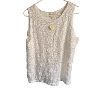 May include: A sleeveless white top featuring an all-over floral lace pattern with a round neckline. The garment is displayed on a white plastic hanger and includes a small, light-colored rectangular tag attached to the front near the collar.