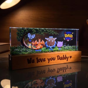 Könnte beinhalten: Rechteckiger, beleuchteter Block mit Holzsockel. Er zeigt eine grüne Landschaft mit Zeichentrickfiguren, einem Baum und einem Schmetterling. Die Worte "Best Dad Ever" und "We love you Daddy" sind sichtbar.