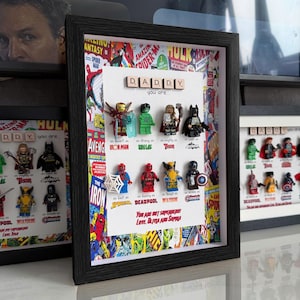 May include: Framed artwork featuring Lego superhero figures, including Iron Man, Hulk, and Batman. The artwork includes the text "Daddy, you are..." and is surrounded by a comic book-themed background. The frame is black.