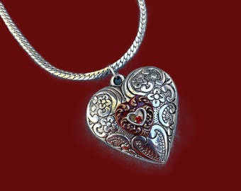 May include: Silver heart pendant necklace with intricate floral and scrollwork designs. The pendant features a smaller heart with a red gemstone. The necklace has a silver chain and is set against a red background.