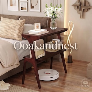 May include: A dark wood side table with a rectangular top and a lower shelf. The top holds a glass vase with white lilies and stacked books. The lower shelf contains a small radio and a woven basket. A white robotic vacuum sits on the floor.