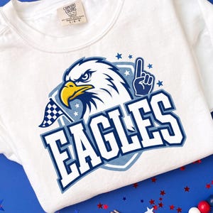 May include: White t-shirt with a graphic of an eagle's head, a yellow beak, and the word "EAGLES" in blue and white. The design includes a checkered flag and a hand with the number one. The shirt has a crew neck and a tag.