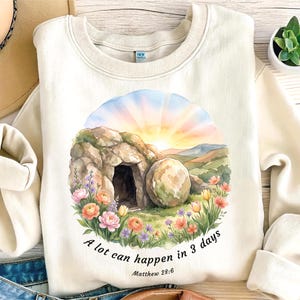 Puede incluir: Sudadera color crema con un diseño de acuarela de una tumba vacía y un sol naciente, rodeada de flores. El texto "A lot can happen in 3 days" y "Matthew 28:6" están impresos debajo. Una prenda superior casual y cómoda.