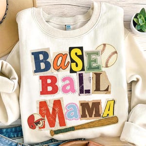 Può includere: Felpa color crema con la scritta "BASEBALL Mama" in stile collage. Il design include una palla da baseball, una mazza e un casco. Le lettere sono in vari colori e caratteri.