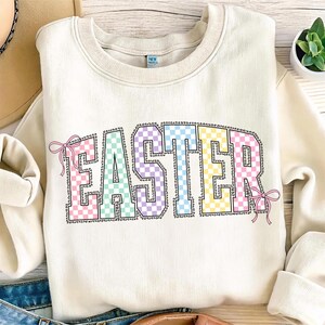 May include: Cream-colored sweatshirt with the word "EASTER" in a colorful checkered pattern. The letters are outlined in black and adorned with pink bows. A casual, festive top for the Easter season.