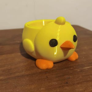 Op de afbeelding: Een felgele kuikenvormige houder met een gladde afwerking. Het figuurtje heeft een kleine oranje snavel, twee oranje voetjes en twee ronde zwarte ogen. Bovenop de kop van het kuiken zit een ronde opening die een holle ruimte creëert.