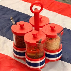 Peut inclure: Un présentoir à épices rouge avec quatre pots en verre transparent, chacun avec un couvercle rouge et une petite cuillère. Le présentoir a une poignée et est décoré de rayures blanches, bleues et rouges. Les pots contiennent diverses épices.