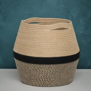 May include: A woven storage basket with a natural beige upper section and a black band separating it from the patterned lower section. The lower section has a black and beige diagonal weave pattern. The basket has two handles.