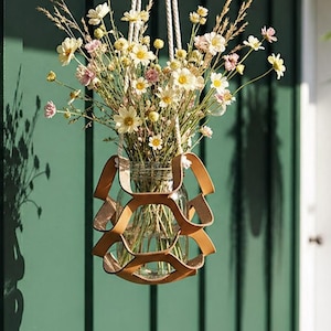 Peut inclure: Un arrangement floral suspendu dans un bocal en verre transparent, suspendu par une corde blanche. Le bocal est maintenu dans un support en cuir marron. L'arrangement comprend des fleurs sauvages et de la verdure. Une fougère suspendue est en arrière-plan.