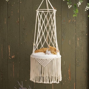 Op de afbeelding: Een crèmekleurig macramé kattenbed hangt aan een wit touw, met een slapende oranje en witte kat erin. Het bed heeft een geweven ruitpatroon en een franjebodem. Een houten krat met lavendel staat op een houten oppervlak.