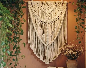 May include: A cream-colored macrame wall hanging with a geometric design is displayed on a terracotta-colored wall. The room is decorated with hanging plants, a wooden side table, and a woven lamp. An open book and a vase of flowers sit on the table.