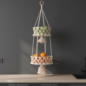 Può includere: Un cestino di frutta in macramè a due livelli, sospeso a un anello di legno. Il cestino superiore contiene mele verdi, mentre il cestino inferiore contiene arance. I cestini sono realizzati in cotone intrecciato color crema, con nappe decorative.