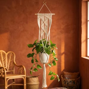 May include: A cream-coloured macrame plant hanger featuring intricate knotted patterns, two small wooden beads, and a long tassel. The hanger supports a white ceramic pot containing a green pothos plant, suspended from a wooden dowel by a cord.