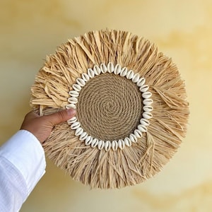 Op de afbeelding: Ronde wanddecoratie van natuurlijke vezels, met een spiraalvormig jute centrum en een ring van kleine witte schelpen. De buitenrand heeft een franje van gedroogd grasachtig materiaal, wat een textuurrijke, bohemien esthetiek creëert.