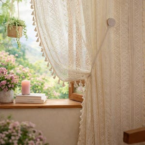Puede incluir: Cortinas de encaje color crema con un patrón delicado y flecos. Una vela rosa encendida está sobre un alféizar de madera con libros y un jarrón con flores rosas. Se ve una cesta colgante con vegetación.