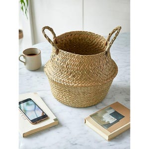 May include: A woven basket with two handles, made of natural-colored straw. The basket sits on a marble surface, next to a mug of coffee, a smartphone, and two books. The basket is round with a flared base.