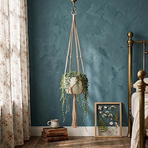 Peut inclure: Un porte-plante en macramé suspendu avec un pot en céramique blanc rempli de plantes vertes retombantes. Le support est suspendu à un crochet de plafond. La pièce a un mur bleu, des rideaux à fleurs et un cadre de lit vintage.