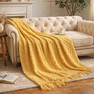 Może przedstawiać: Musztardowy, dziergany koc z rombowym wzorem i frędzlami, narzucony na beżową, pikowaną sofę. Koc jest wykonany z miękkiego materiału. Na sofie stoi filiżanka ze spodkiem, co dodaje przytulności.