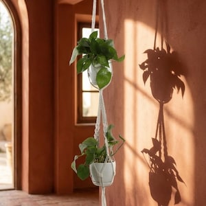 May include: A white macrame plant hanger featuring two tiers, each holding a white ceramic pot with a green leafy plant. The hanger is suspended from a metal hook and includes a long, decorative fringe tassel at the bottom end.