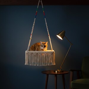 May include: A hanging cat bed made of woven macrame with a fringed bottom. The bed is suspended from a wooden beam by two white ropes with colorful beads. An orange tabby cat is resting inside the bed.