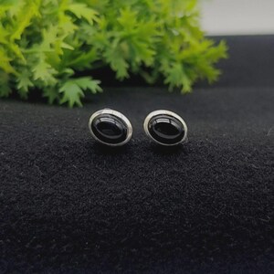 Puede incluir: Un par de pendientes de botón con piedras negras ovaladas y lisas engastadas en monturas de metal plateado pulido. Los pendientes tienen un diseño sencillo y redondeado que resalta el contraste entre el centro oscuro y brillante y el borde metálico.