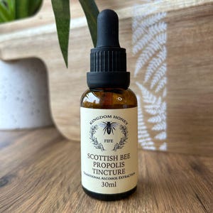 May include: A brown glass bottle with a black dropper. The label reads "Kingdom Honey Fife Scottish Bee Propolis Tincture Traditional Alcohol Extraction 30ml" with a bee illustration. The bottle is on a wooden surface.