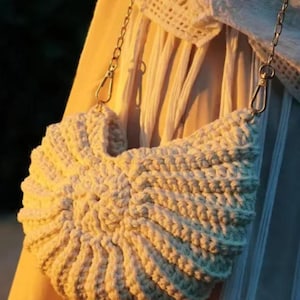May include: A cream-colored crochet handbag with a distinct shell-like, fan-shaped design. The bag features a thick, textured yarn construction and is attached to a metallic gold-toned chain strap with lobster claw clasps on both sides.
