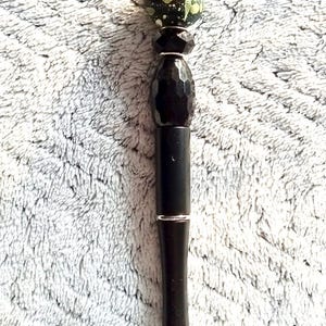 May include: A black pen with a decorative design. The pen has a black barrel with a silver band, a faceted black section, and a green and black speckled top. The pen has a silver tip.