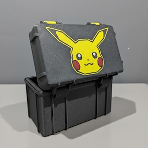May include: A black storage case featuring a yellow Pikachu design on the lid. The case has a rectangular shape with a hinged lid and yellow details. The lid is open, showing the inside of the case.
