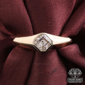 Peut inclure: Une bague en or avec une pierre précieuse carrée et transparente sertie dans une lunette. Le corps de la bague est en or lisse et poli. La bague est présentée sur un tissu rouge foncé, créant un contraste.
