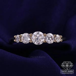 Puede incluir: Un anillo de oro con cinco piedras redondas transparentes dispuestas en fila, con la piedra más grande en el centro. La banda es fina y metálica, sujetando las piedras en un engaste de garras. El texto FACET.GURU HANDCRAFTED es visible.