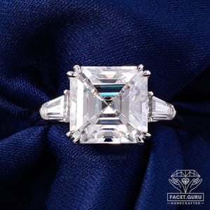 May include: A silver ring featuring a large, square-cut, clear gemstone. Two smaller, rectangular gemstones are set either side of the main stone. The ring is displayed on a dark blue fabric.