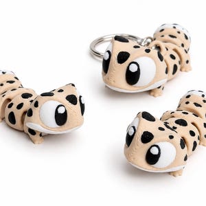 Op de afbeelding: Drie tan en zwart gevlekte gecko beeldjes. Eén is bevestigd aan een zilveren sleutelhanger. De gekko's hebben grote witte ogen met zwarte pupillen. De gekko's staan op een witte achtergrond.