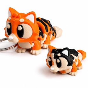 Op de afbeelding: Twee 3D-geprinte kattenbeeldjes. Eén is een sleutelhanger met een zilveren ring, de andere is een kleinere versie. Beide zijn oranje met zwarte strepen, witte accenten en grote zwarte ogen. De kattenbeeldjes zijn ontworpen in een cartoonstijl.