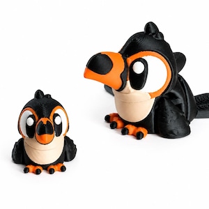 Op de afbeelding: Twee 3D-geprinte vogelbeeldjes. De vogels zijn zwart met oranje snavels en poten en crèmekleurige borst. De grotere vogel ligt, terwijl de kleinere vogel rechtop staat. Beide hebben grote witte ogen met zwarte pupillen.