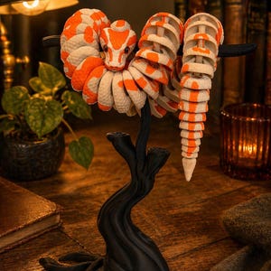 Può includere: Una scultura decorativa a forma di serpente arancione e bianco, avvolta attorno a un supporto nero a forma di albero. Il corpo segmentato e la testa del serpente sono dettagliati. La scultura è esposta su una superficie di legno.