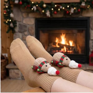 May include: Tan socks with two crocheted snowman ornaments. The snowmen have tan bodies, red and green scarves, green button eyes, and white and red carrot noses. A fireplace with a lit fire is in the background.