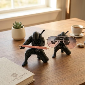 Może przedstawiać: Dwa czarne organizery na biurko ninja, jeden trzymający różowy długopis, a drugi okulary przeciwsłoneczne typu aviator. Mały sukulent w białej doniczce i książka z napisem "NINJA DESKWARES EST. 2023" również znajdują się na drewnianym stole.