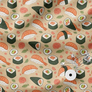 May include: Beige fabric featuring a repeating pattern of sushi rolls, nigiri, and temaki. The design includes pink ginger, green leaves, and pink flower accents. Safety pins and a white button are also visible.