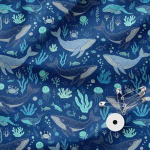 Può includere: Un tessuto blu scuro con un motivo ripetuto di balene, meduse, granchi e alghe azzurre. Un piccolo oggetto circolare bianco e diverse spille da balia in metallo argentato sono posizionati sul lato inferiore destro del tessuto.