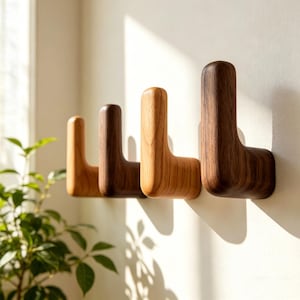 Puede incluir: Cuatro perchas de pared de madera en tonos marrones variados están montadas en una pared blanca. Los ganchos en forma de L tienen una parte superior redondeada y una base plana. La veta de la madera es visible.