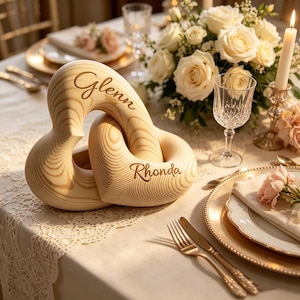 May include: A light-coloured wooden sculpture featuring two interlocking heart shapes. The top heart is engraved with the name Glenn, and the bottom heart is engraved with the name Rhonda. The wood grain is visible throughout the smooth, curved surface.