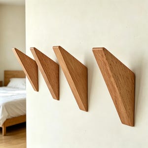 May include: Four wooden wall hooks with a triangular design are mounted on a white wall. The hooks are made of light-coloured wood with visible grain. The hooks are angled, creating a modern and minimalist aesthetic.