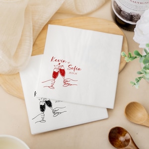 May include: White paper napkins with red and black illustrations. One napkin features the names "Kevin + Sofia" and the date "2007.26" with two champagne glasses clinking. The other napkin has a similar design in black.